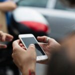 Close-up of hands holding a white smartphone as a group gathers around in the background, out of focus.