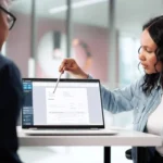 Woman in a denim shirt points at a laptop screen during a meeting with a man sitting opposite her.