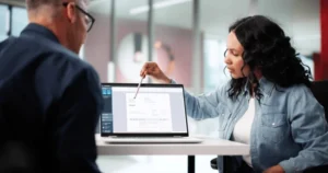 Woman in a denim shirt points at a laptop screen during a meeting with a man sitting opposite her.