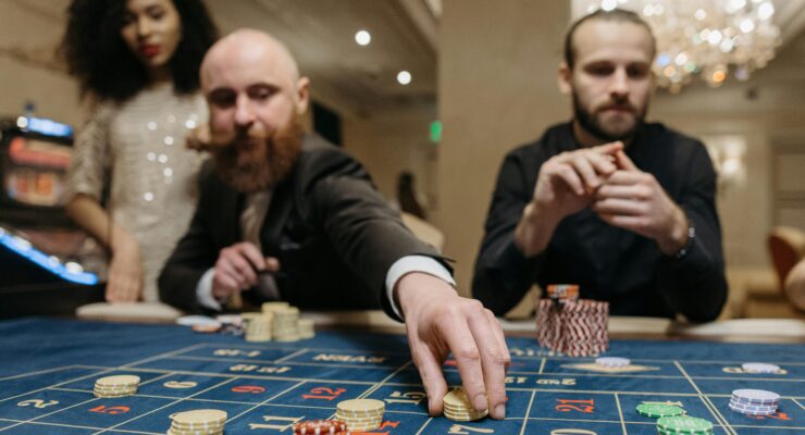 Close-up of a casino roulette table with a player's hand stacking yellow chips on a bet layout; other players are blurred in the background.