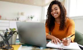 Woman with curly hair wearing an orange shirt, typing at a laptop on a bright desk.