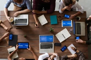 Top-down view of a diverse team around a wooden table with laptops, tablets, notebooks, and coffee cups collaborating.