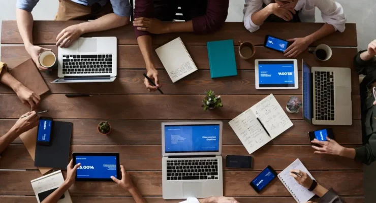 Top-down view of a diverse team around a wooden table with laptops, tablets, notebooks, and coffee cups collaborating.